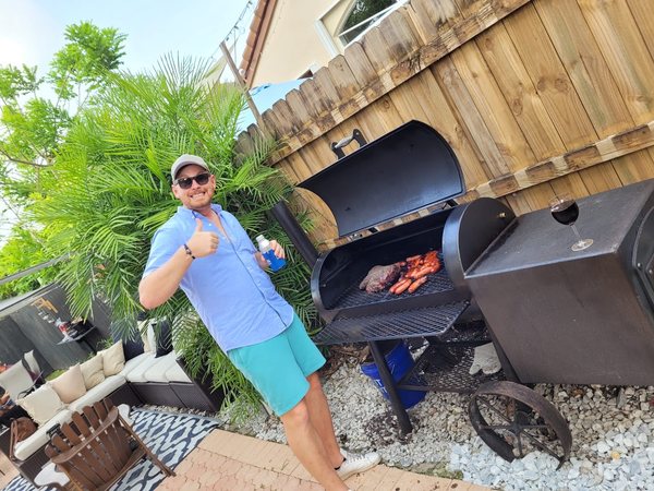 Andreas giving a thumbs up beside the offset smoker in a Miami backyard, sausages and a brisket on the grate