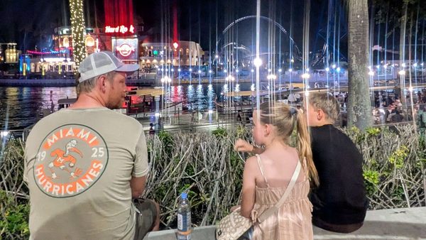 Andreas, Olivia and Jayden sitting by the water at night with lights and a roller coaster behind